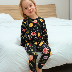 A child sitting on a bed wearing a long-sleeved black pajama set with a colorful floral pattern.